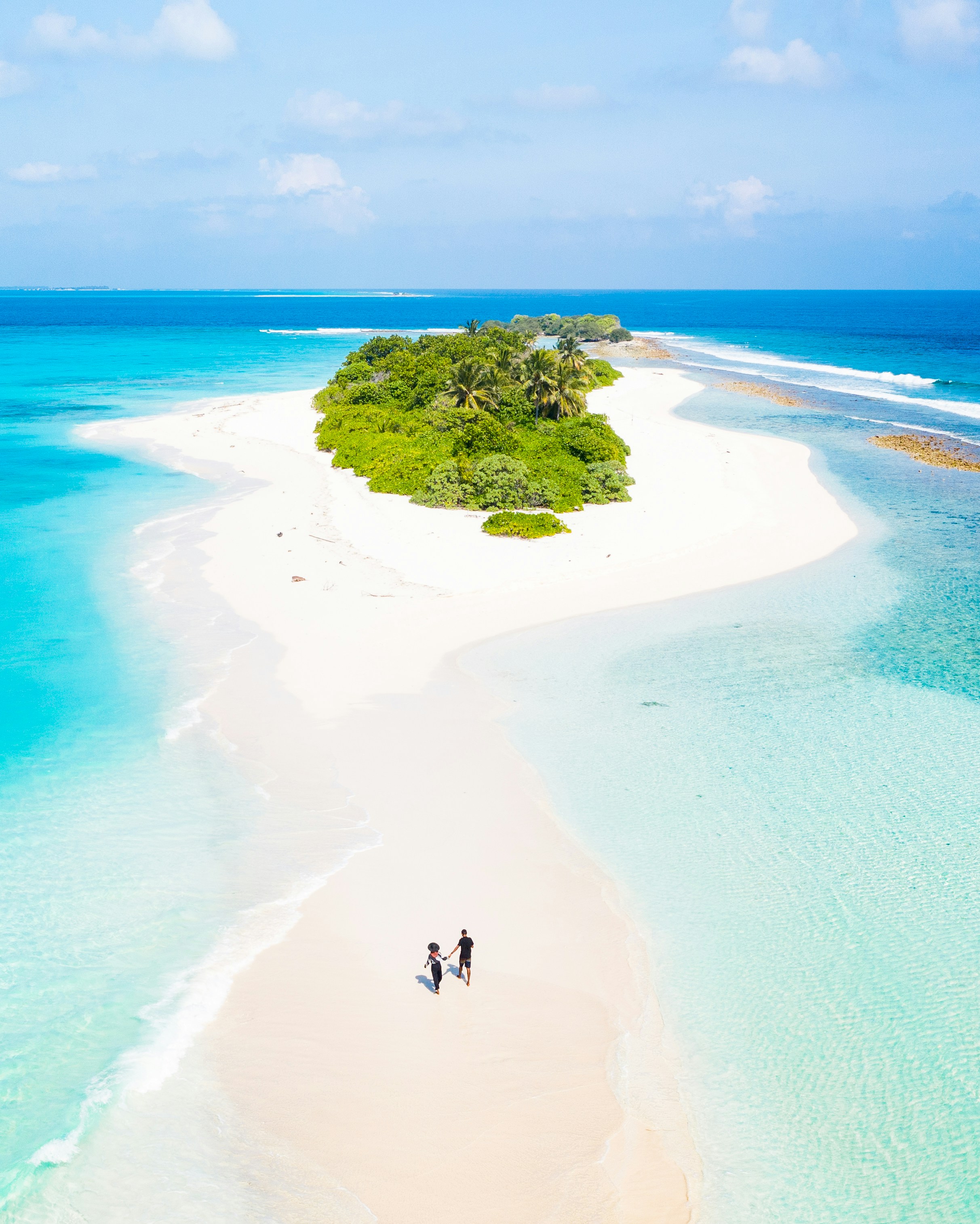 Aerial view of a couple walking hand in hand along a white sandbar leading to a small tropical island surrounded by turquoise water